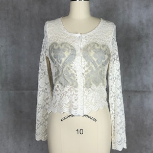 Puede incluir: Un cárdigan de encaje blanco con un dobladillo festoneado y mangas largas. El cárdigan tiene cierre de botones y presenta un delicado patrón floral con un diseño en forma de corazón más grande en el centro.