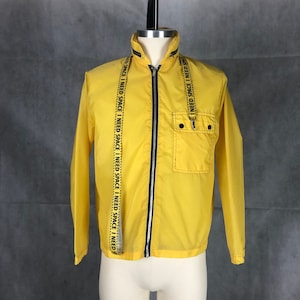 May include: A yellow nylon jacket with a black zipper and a black and yellow strap with the text "I NEED SPACE I NEED SPACE I NEED SPACE I NEED SPACE I NEED SPACE I NEED" repeated. The jacket has a black zipper and a black pocket with a button closure.