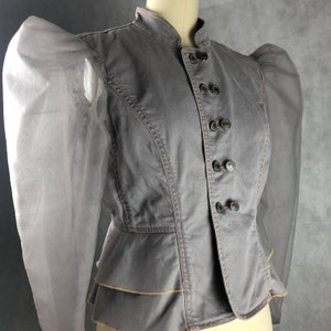 K&ouml;nnte beinhalten: Eine graue Jeansjacke mit gerafftem Saum und durchsichtigen, bauschigen &Auml;rmeln. Die Jacke hat einen Stehkragen und einen Knopfverschluss.