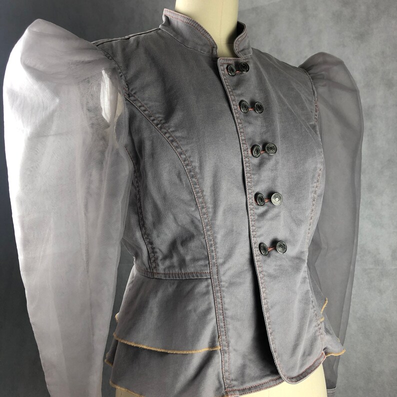 K&ouml;nnte beinhalten: Eine graue Jeansjacke mit gerafftem Saum und durchsichtigen, bauschigen &Auml;rmeln. Die Jacke hat einen Stehkragen und einen Knopfverschluss.