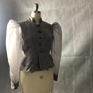 K&ouml;nnte beinhalten: Eine graue, taillierte Jacke mit einem Stehkragen und einer Reihe von Kn&ouml;pfen vorne. Die Jacke hat lange &Auml;rmel mit durchsichtigen, bauschigen &Auml;rmeln.