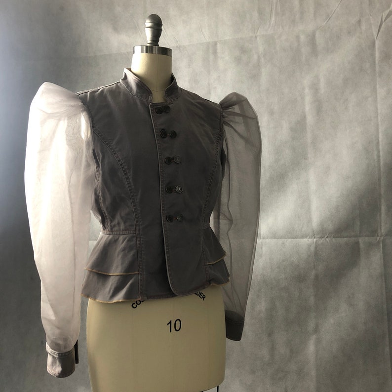 K&ouml;nnte beinhalten: Eine graue, taillierte Jacke mit einem Stehkragen und einer Reihe von Kn&ouml;pfen vorne. Die Jacke hat lange &Auml;rmel mit durchsichtigen, bauschigen &Auml;rmeln.