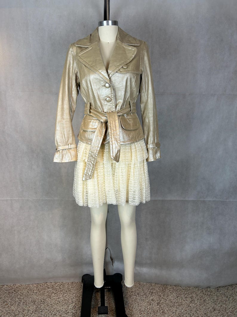 May include: A gold metallic jacket with a belt and a white lace skirt. The jacket has a button closure and a collar.