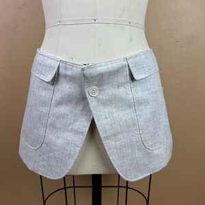 May include: A light gray, tailored garment with two front pockets and a button closure. The garment is displayed on a mannequin and appears to be made of a textured fabric. The design features a curved hemline.