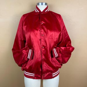 May include: A bright red satin varsity jacket featuring a snap-button front closure, long sleeves with ribbed cuffs, and a ribbed collar and hem. The collar, cuffs, and hem are accented with two thin white stripes against the red fabric.