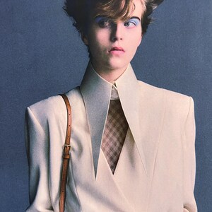 May include: A cream-colored double-breasted blazer with sharp, elongated lapels and structured shoulders. The blazer is worn over a brown and white checkered shirt. A thin, tan leather shoulder strap with a gold buckle is visible on the right side.