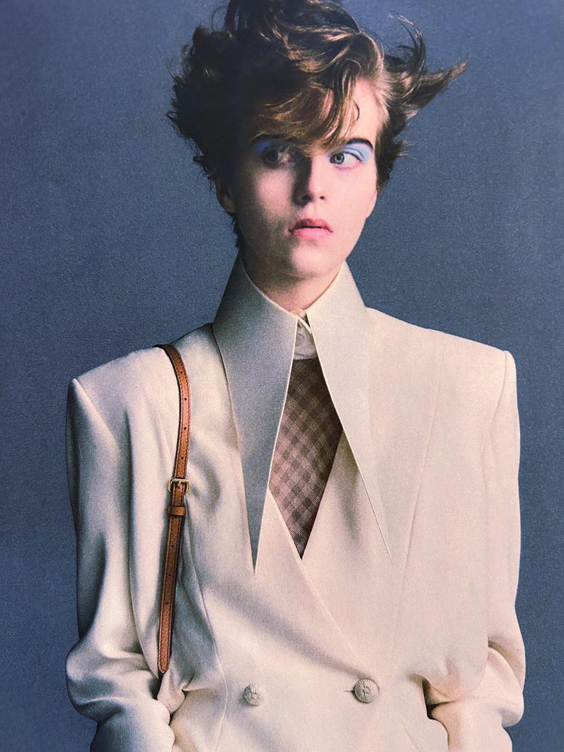 May include: A cream-colored double-breasted blazer with sharp, elongated lapels and structured shoulders. The blazer is worn over a brown and white checkered shirt. A thin, tan leather shoulder strap with a gold buckle is visible on the right side.