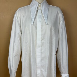 May include: A long-sleeved white dress shirt featuring a prominent, oversized pointed collar and a vertical pleated bib front. The shirt includes a concealed button placket, long sleeves with wide cuffs, and a smooth, solid white fabric construction.