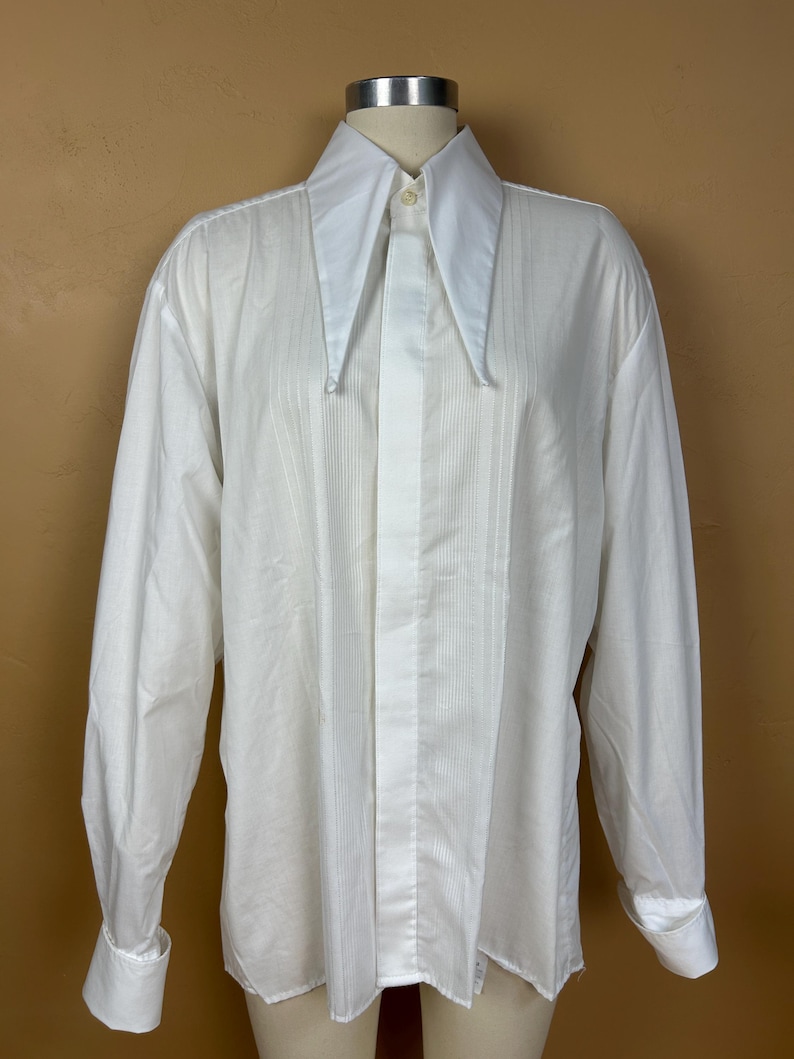 May include: A long-sleeved white dress shirt featuring a prominent, oversized pointed collar and a vertical pleated bib front. The shirt includes a concealed button placket, long sleeves with wide cuffs, and a smooth, solid white fabric construction.