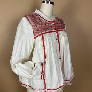 May include: White long-sleeve blouse with colorful embroidered yoke and red trim. The blouse features a button-down front and frayed edges. The embroidery includes pink, red, and blue threads. The sleeves are long and loose.
