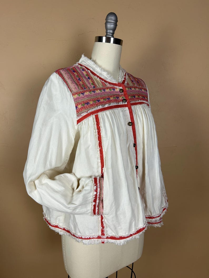 May include: White long-sleeve blouse with colorful embroidered yoke and red trim. The blouse features a button-down front and frayed edges. The embroidery includes pink, red, and blue threads. The sleeves are long and loose.