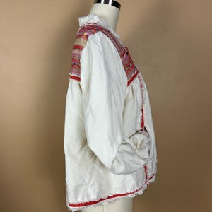 May include: White jacket with colorful embroidered shoulder and neckline details. The jacket has long sleeves and red trim along the edges and cuffs. The fabric has a slightly frayed texture, adding a unique touch to the garment.