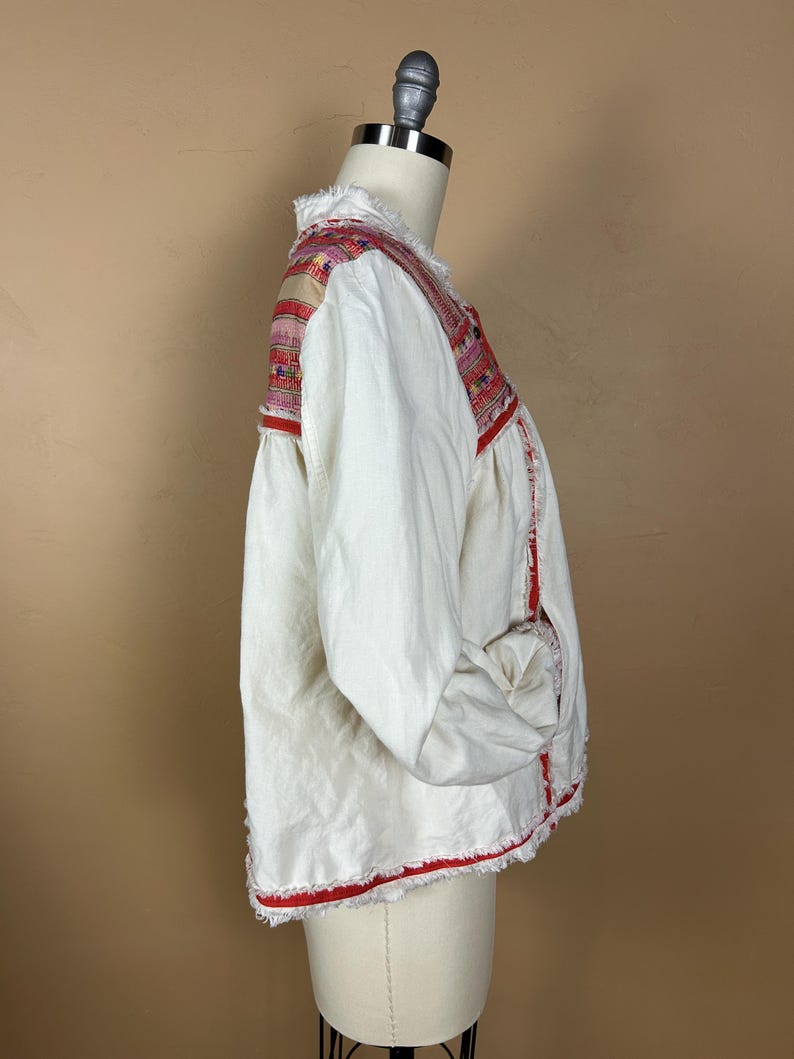 May include: White jacket with colorful embroidered shoulder and neckline details. The jacket has long sleeves and red trim along the edges and cuffs. The fabric has a slightly frayed texture, adding a unique touch to the garment.