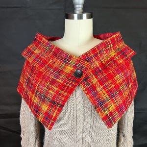May include: A red plaid collar with yellow, purple, and black accents, fastened with a black button. The collar is displayed on a mannequin over a light gray sweater with a cable knit design.