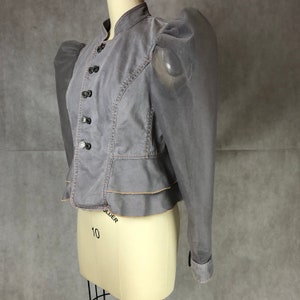 K&ouml;nnte beinhalten: Eine graue, kurze Jacke mit einem R&uuml;schen-Saum und durchsichtigen, bauschigen &Auml;rmeln. Die Jacke hat einen Stehkragen und eine Reihe von Kn&ouml;pfen an der Vorderseite.