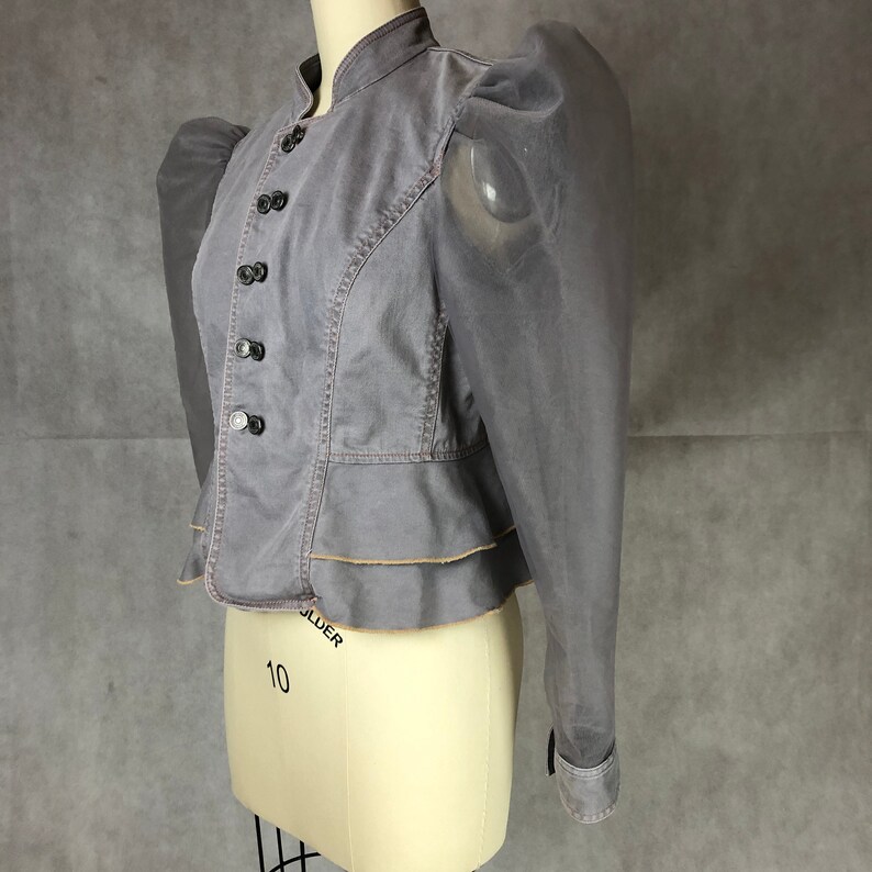 K&ouml;nnte beinhalten: Eine graue, kurze Jacke mit einem R&uuml;schen-Saum und durchsichtigen, bauschigen &Auml;rmeln. Die Jacke hat einen Stehkragen und eine Reihe von Kn&ouml;pfen an der Vorderseite.