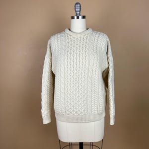 May include: Cream-colored cable knit sweater displayed on a mannequin. The sweater features a crew neck and intricate cable patterns across the front, sleeves, and back. The garment is made of a thick, textured material, suggesting warmth and comfort.