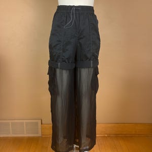 May include: Black cargo pants with a drawstring waist and a sheer, flowing lower half. The pants have pockets on the sides and are cuffed at the knee. The fabric appears to be lightweight and suitable for warmer weather.
