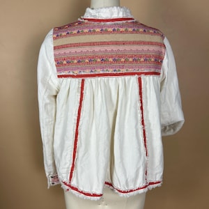 May include: White long-sleeved blouse with colorful embroidered yoke and red trim. The blouse features a ruffled collar and a loose, flowing fit. The embroidery includes pink, red, green, and blue threads.