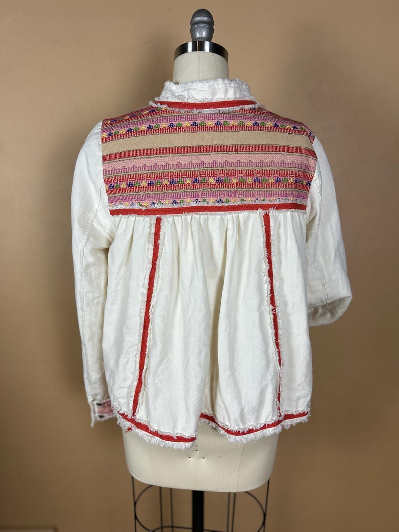 May include: White long-sleeved blouse with colorful embroidered yoke and red trim. The blouse features a ruffled collar and a loose, flowing fit. The embroidery includes pink, red, green, and blue threads.