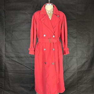 May include: A bright red trench coat with a double-breasted design, featuring a belt, and button details. The coat has long sleeves and a classic collar. The coat is displayed on a mannequin.