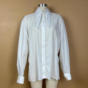 May include: A crisp white long-sleeve button-up shirt featuring a prominent pointed collar and a vertical pleated bib front. The garment includes long sleeves with wide cuffs and a central button placket, presented on a mannequin torso.