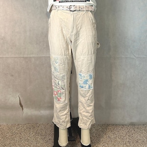 May include: Beige utility trousers featuring a light-coloured belt with a floral pattern. The trousers have multiple patchwork squares in shades of blue, pink, and white sewn onto the front of the legs, along with a white t-shirt visible at the waist.