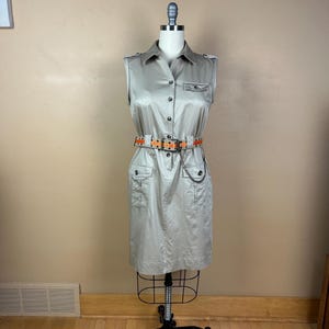 May include: A sleeveless beige button-down dress featuring a collared neckline, two chest pockets, and two lower front pockets. The dress is cinched at the waist with a belt that has an orange patterned strap and a silver chain detail attached.