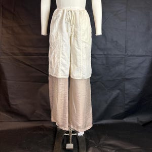 Puede incluir: Pantalones de dos capas en un maniquí. La capa superior es de tela acolchada color crema con cintura con cordón. La capa inferior es de material transparente marrón claro, creando una silueta única y fluida. Los pantalones se exhiben en un maniquí.