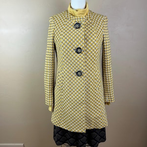 May include: A long-sleeved coat featuring a yellow, white, and black houndstooth pattern. The coat has a high collar, three large dark buttons down the center front, and is layered over a yellow turtleneck top and a dark patterned skirt.