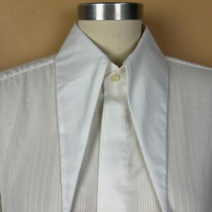 May include: A crisp white button-down shirt featuring an elongated, sharp-pointed collar and a vertical pleated front placket. The garment is displayed on a mannequin, showcasing the smooth fabric texture and the single visible top button.