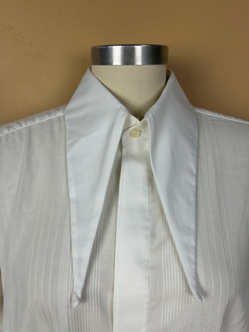May include: A crisp white button-down shirt featuring an elongated, sharp-pointed collar and a vertical pleated front placket. The garment is displayed on a mannequin, showcasing the smooth fabric texture and the single visible top button.