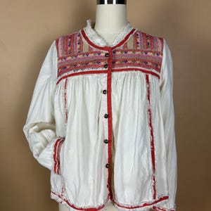May include: White long-sleeve shirt with red trim and embroidered details. The shirt features a button-up front and decorative embroidery across the chest and sleeves. The shirt has a relaxed fit and frayed edges.