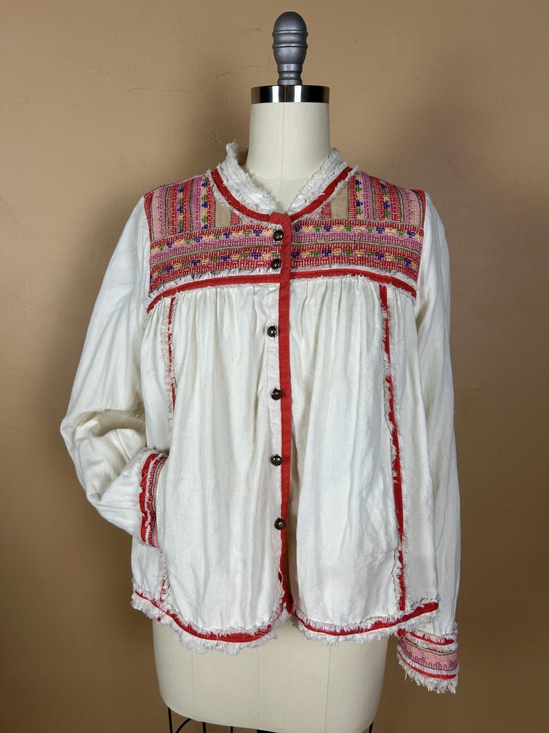 May include: White long-sleeve shirt with red trim and embroidered details. The shirt features a button-up front and decorative embroidery across the chest and sleeves. The shirt has a relaxed fit and frayed edges.