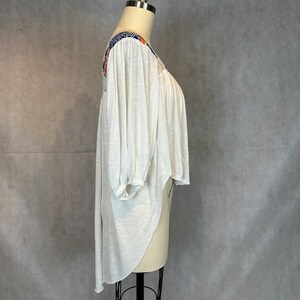 Puede incluir: Una blusa blanca, ligera, de manga larga con cuello redondo alto y dobladillo asim&eacute;trico. La blusa tiene un estampado floral colorido en los hombros y las mangas.