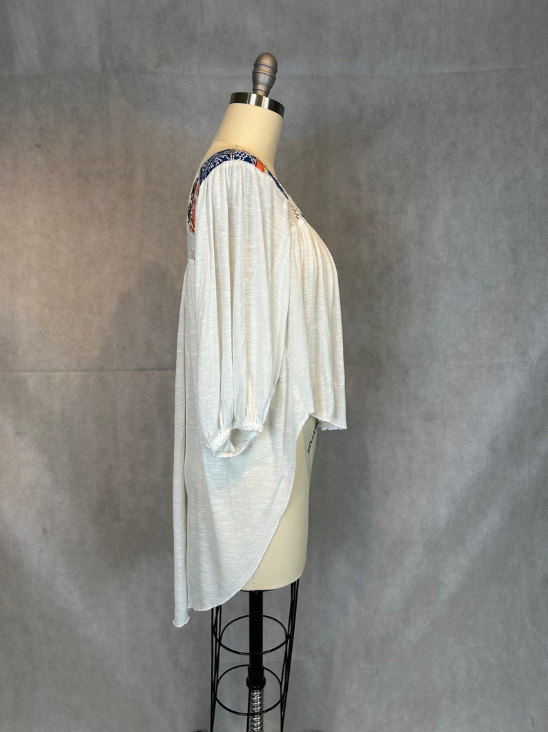 Puede incluir: Una blusa blanca, ligera, de manga larga con cuello redondo alto y dobladillo asim&eacute;trico. La blusa tiene un estampado floral colorido en los hombros y las mangas.