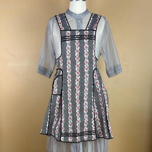 May include: A vintage-style apron dress with a black and white checkered pattern and vertical floral stripes. The dress features a square neckline, black trim, and pockets. A sheer gray blouse is worn underneath, creating a layered effect.