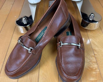 May include: A pair of brown leather loafers with silver-colored metal accents. The shoes have a classic design with a rounded toe and a slip-on style. The loafers are displayed on a wooden surface.