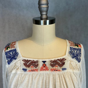Puede incluir: Blusa blanca de algod&oacute;n con cuello cuadrado y patrones geom&eacute;tricos bordados en azul, naranja y rojo.