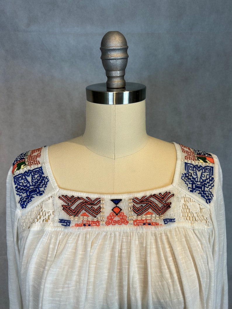 Puede incluir: Blusa blanca de algod&oacute;n con cuello cuadrado y patrones geom&eacute;tricos bordados en azul, naranja y rojo.