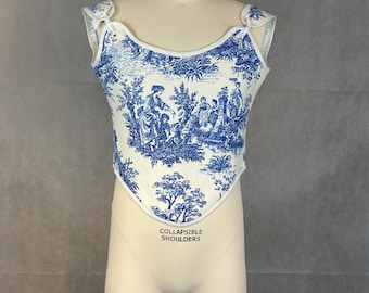 May include: A cream-colored corset with a blue toile pattern featuring figures and landscapes. The corset has a curved neckline and white trim. The words "COLLAPSIBLE SHOULDERS" are printed on the front.