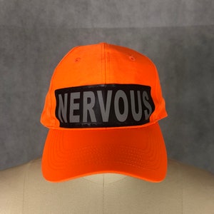 May include: An orange baseball cap with a black reflective band that says "NERVOUS" in white letters.