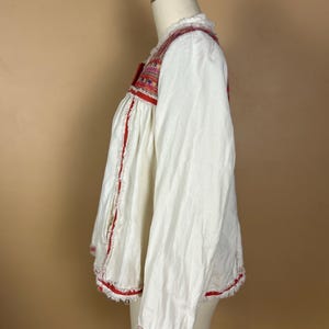 May include: White long-sleeved blouse with red and multicolored embroidered details. The blouse features a square neckline and is trimmed with red fringe along the edges. The sleeves have embroidered cuffs.