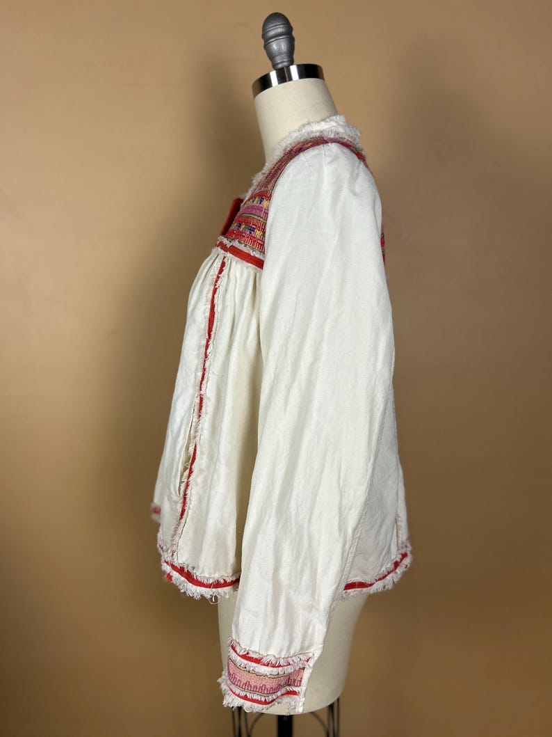 May include: White long-sleeved blouse with red and multicolored embroidered details. The blouse features a square neckline and is trimmed with red fringe along the edges. The sleeves have embroidered cuffs.