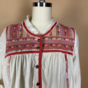 May include: White blouse with a colorful embroidered yoke. The yoke features a geometric pattern in red, pink, and blue. The blouse has a ruffled collar and red trim with wooden buttons.