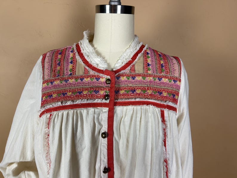 May include: White blouse with a colorful embroidered yoke. The yoke features a geometric pattern in red, pink, and blue. The blouse has a ruffled collar and red trim with wooden buttons.