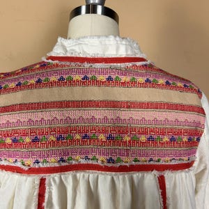 May include: A white blouse with a detailed embroidered yoke. The yoke features a beige center panel with horizontal bands of red, pink, and colorful geometric patterns. The neckline has a ruffled trim, and the blouse has a loose fit.