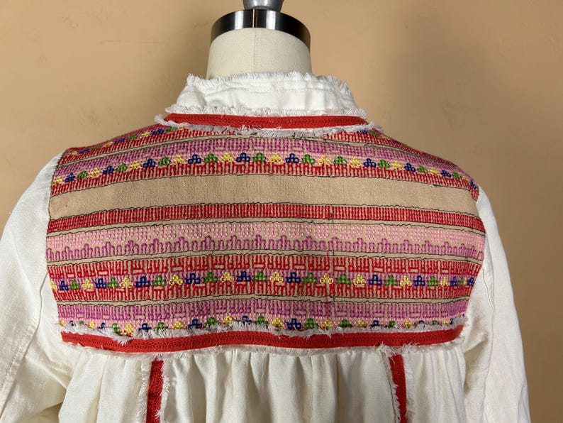 May include: A white blouse with a detailed embroidered yoke. The yoke features a beige center panel with horizontal bands of red, pink, and colorful geometric patterns. The neckline has a ruffled trim, and the blouse has a loose fit.