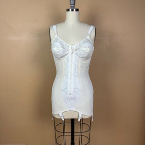 Puede incluir: Una faja moldeadora blanca de cuerpo completo con cierre de cremallera frontal, copas con detalles de encaje y tirantes ajustables. La prenda incluye un panel central de encaje vertical y cuatro ligueros sujetos en el dobladillo inferior.