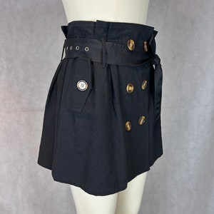 Puede incluir: Una falda negra de cintura alta con un cinturón negro ancho y botones dorados. La falda tiene un cierre de doble botonadura y un diseño plisado.